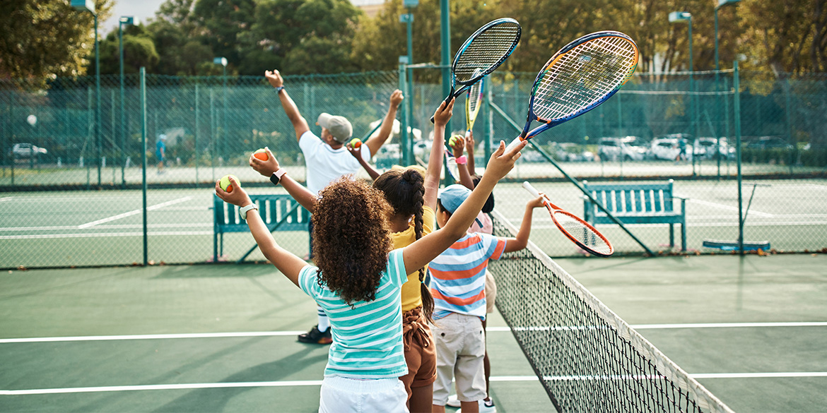 Youth players holding tennis balls and racquets in the air getting trained on how to serve by a coach.