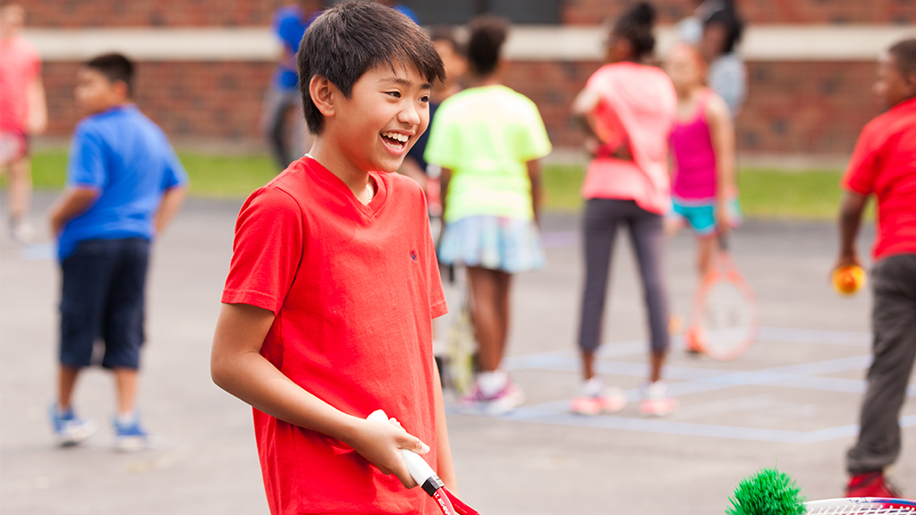 Young male tennis player smiling with racquet in hand and other children in background.