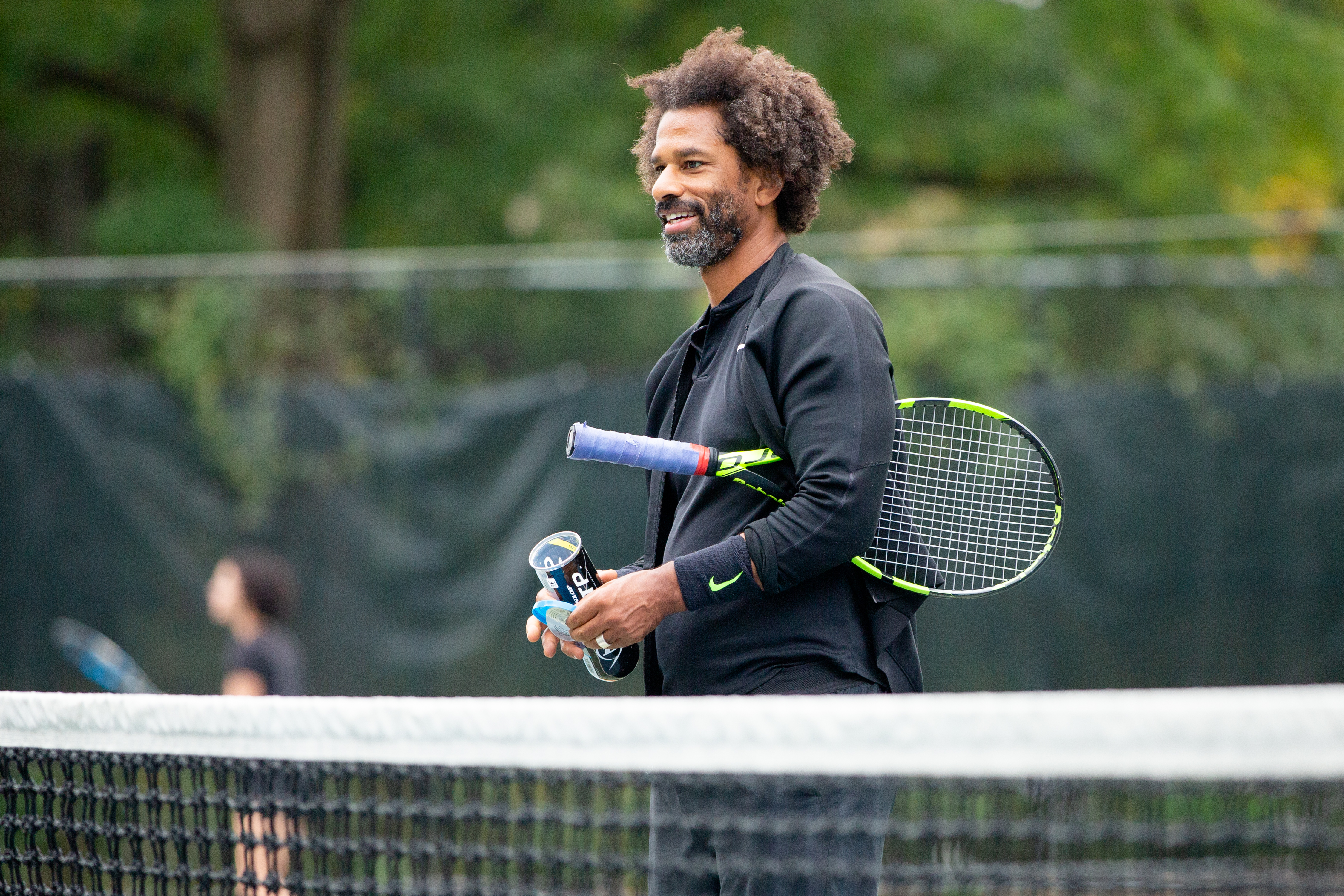 A man smiling in black long sleeve shirt standing behind a net, holding a racquet under his left arm and  a can of tennis balls in his hands. 