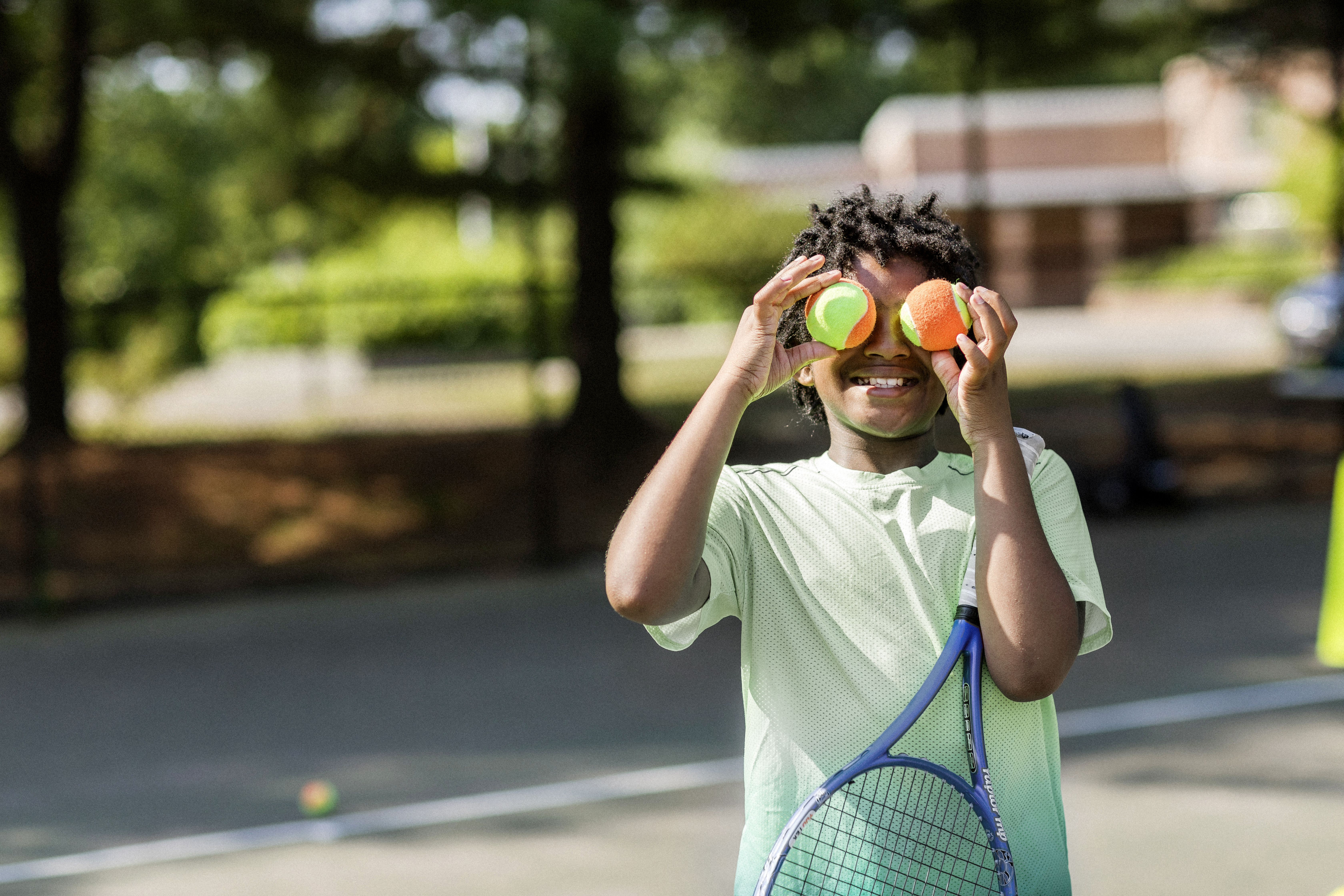 Young boy with a racquet tucked under his arm smiles at the camera while holding two orange and green tennis balls over each of his eyes.
