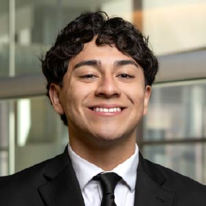 Erick Diaz smiling at the camera while wearing a black suit, white shirt and black tie.