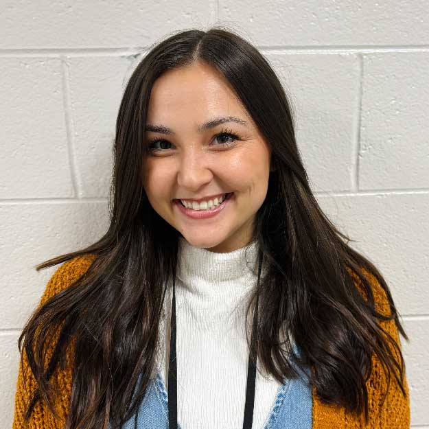 Kennedy Broadwell smiling with long dark hair wearing a white sweater and mustard-colored cardigan against a plain white wall.