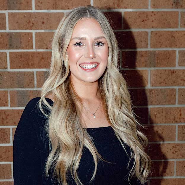 Kaylee Dean smiling indoors in front of a brick background.