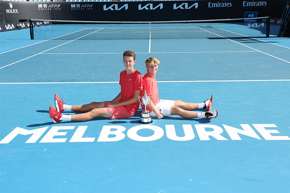 Dos niños se sientan en una cancha de tenis con su trofeo.